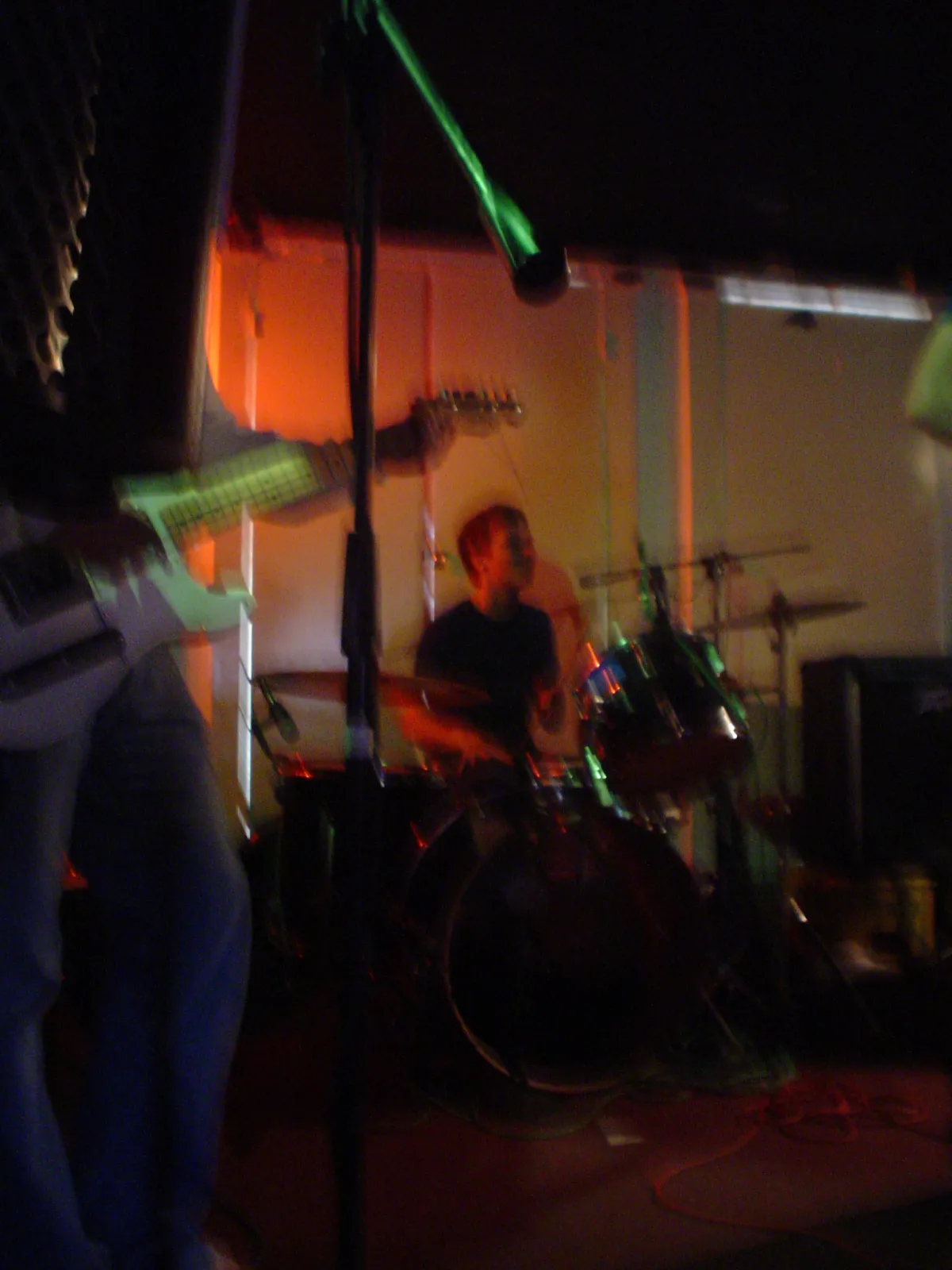 A drummer behind his kit on a dark stage, seen past a guitarist in the foreground.