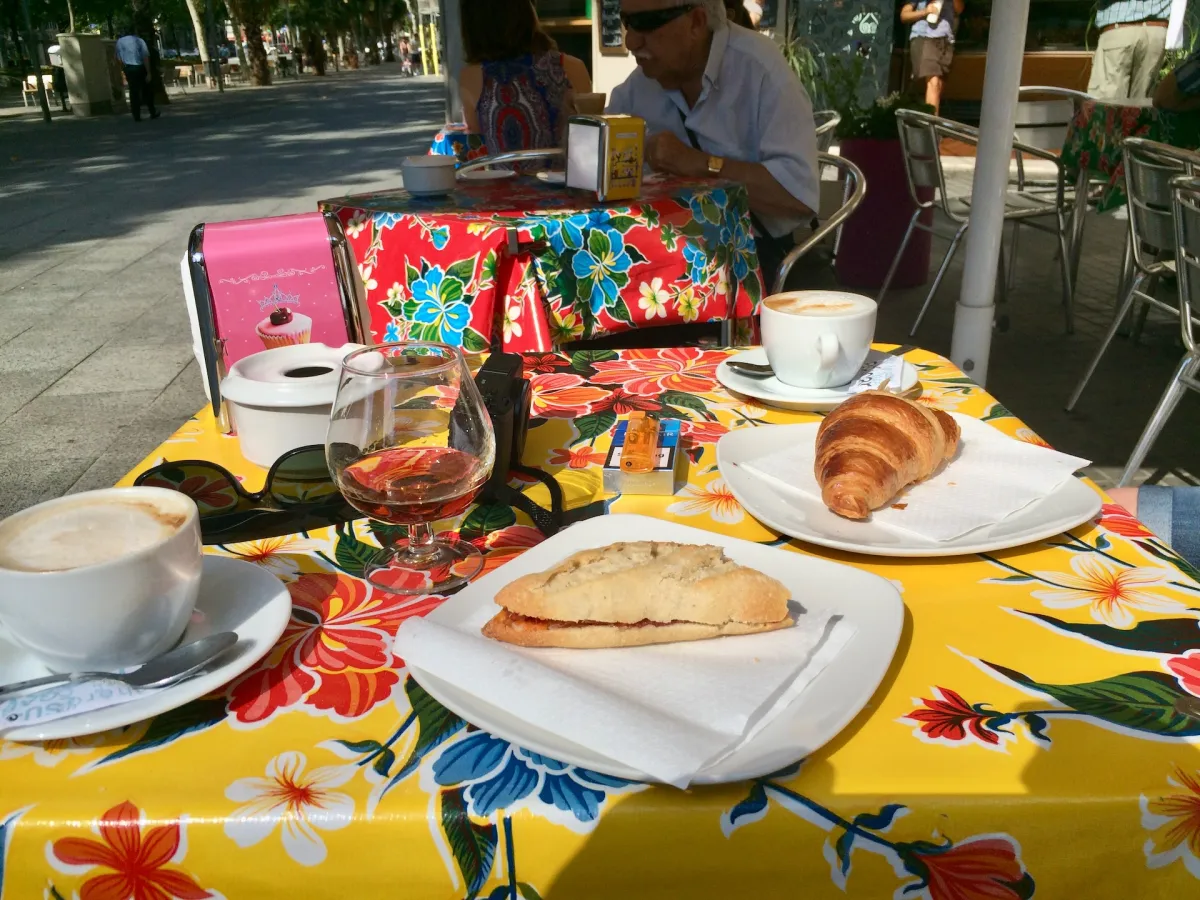 A café table with coffee, a croissant, a pastry and a glass of wine on a floral tablecloth.
