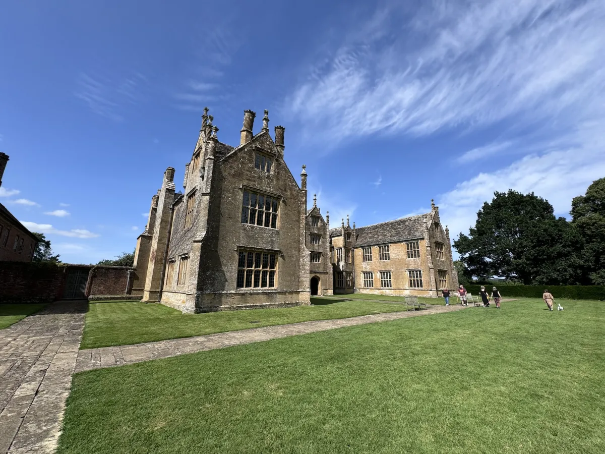 A Tudor manor house of honey-coloured stone, with a lawn in front.