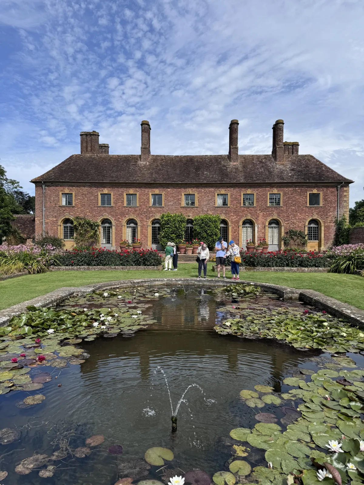 A brick house behind a lily pond with a fountain, and visitors on the path.
