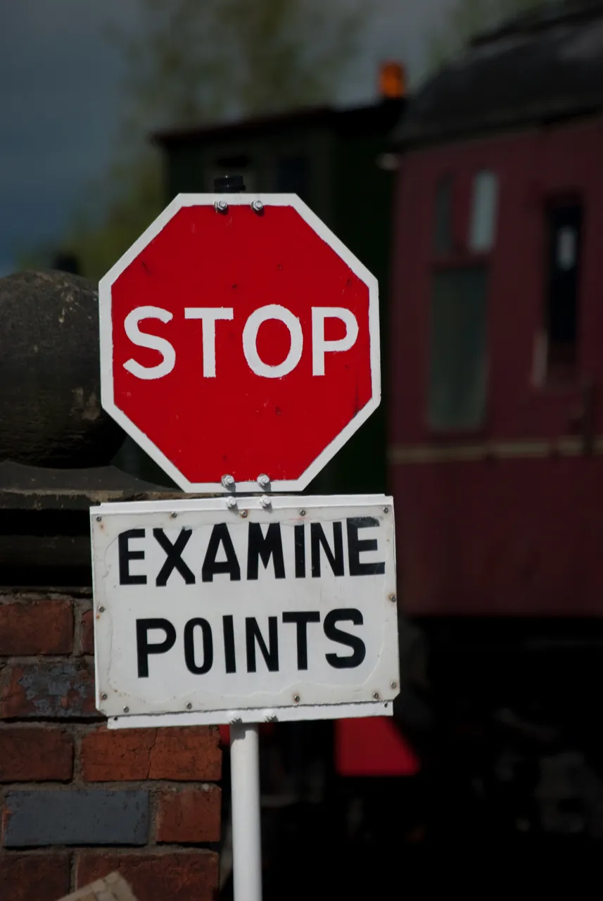 A red stop sign above a plate reading Examine Points, with railway carriages behind.