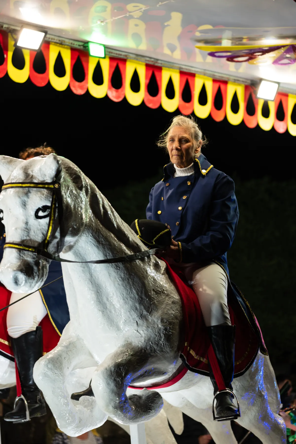A performer in a blue riding jacket on a model white horse, beneath a lit canopy.