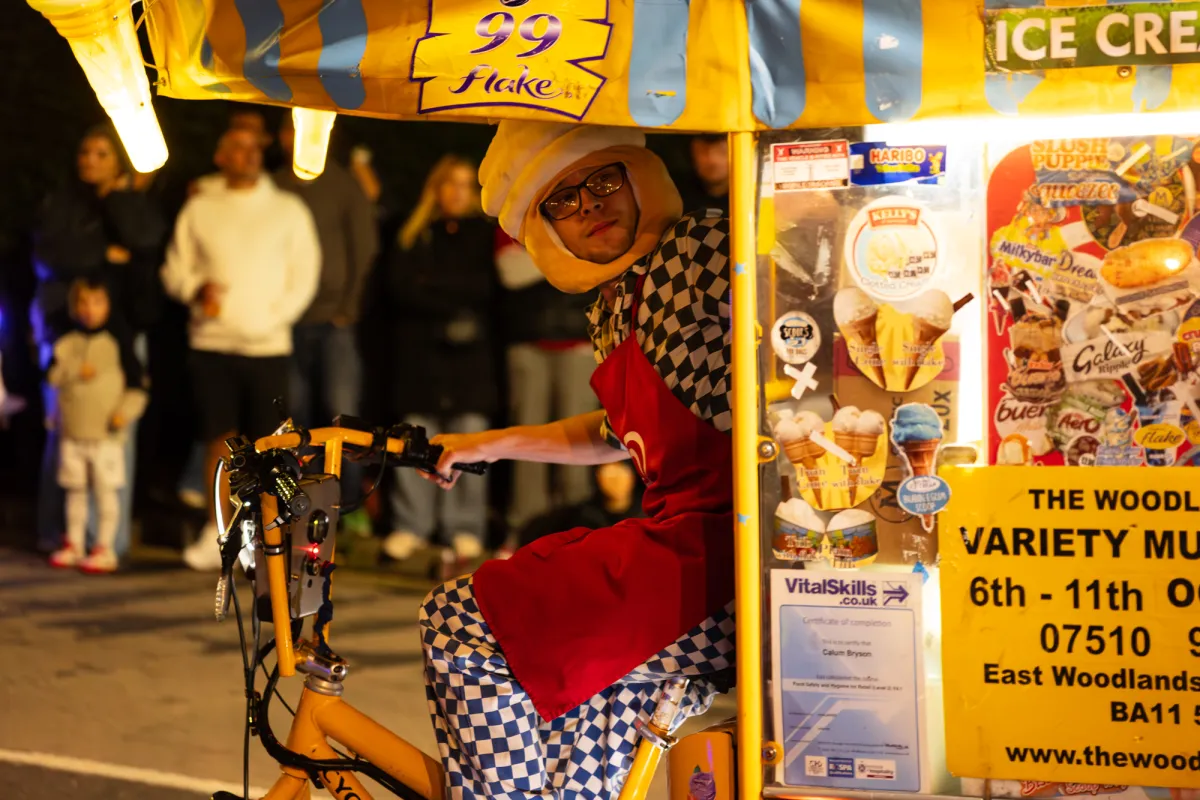A performer in a chequered costume riding a bicycle beside an ice cream stall.
