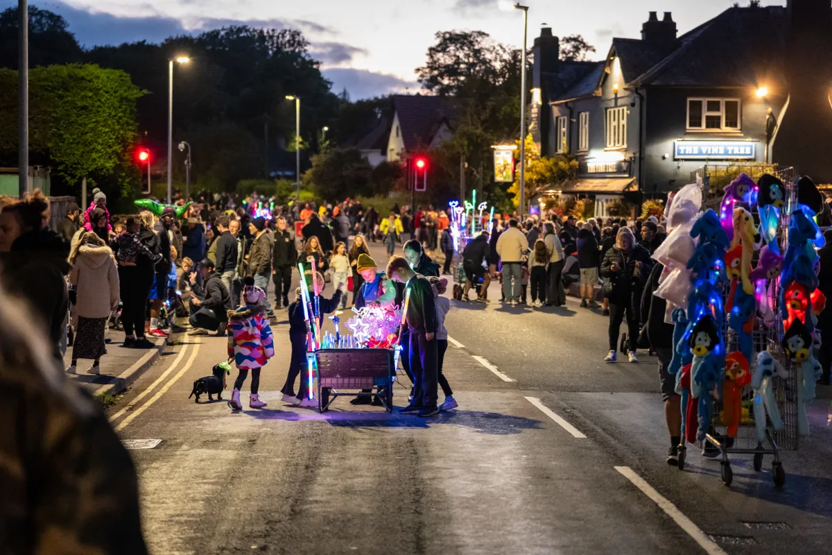 A crowd lining a street at night, waiting for a carnival procession to pass.