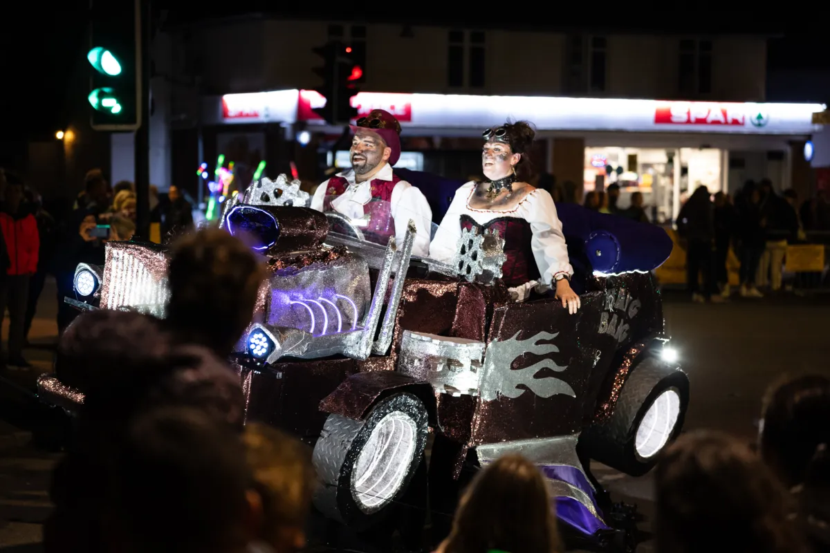 Two performers riding a decorated vintage car through the crowd at night.