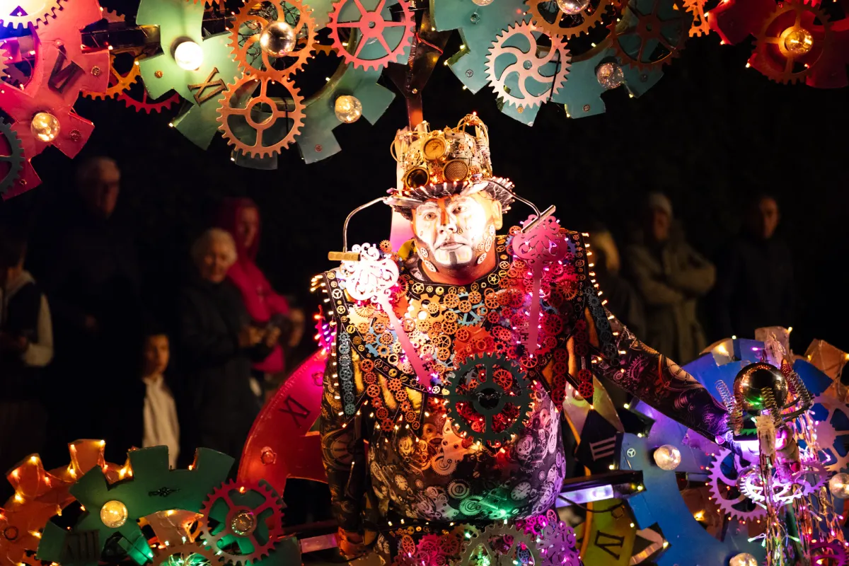 A performer in a crown and beaded costume, surrounded by painted cogs.