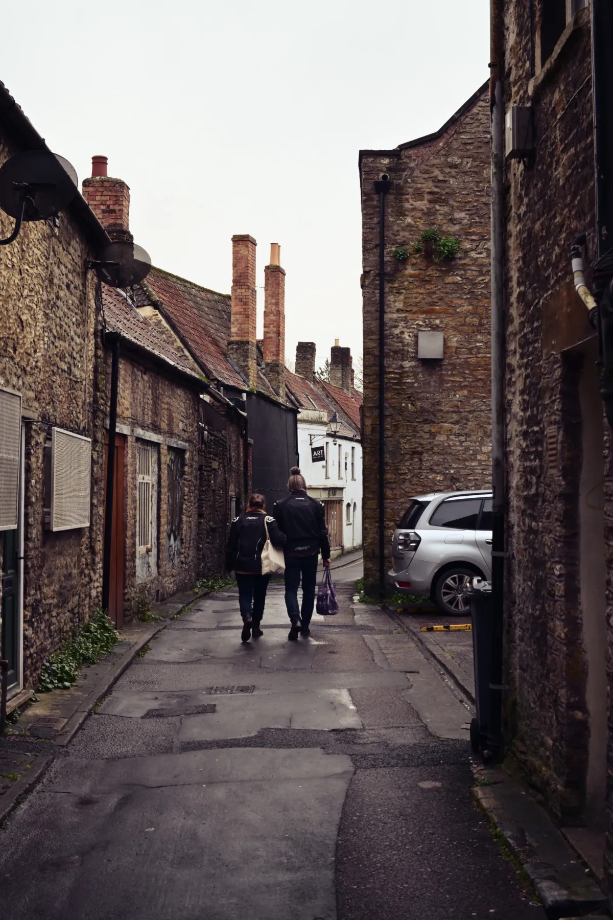 Two people walking away down a narrow lane between stone buildings.