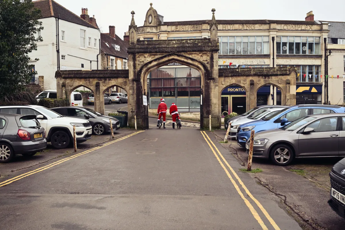 Two cyclists in Father Christmas suits riding through a stone archway in a car park.