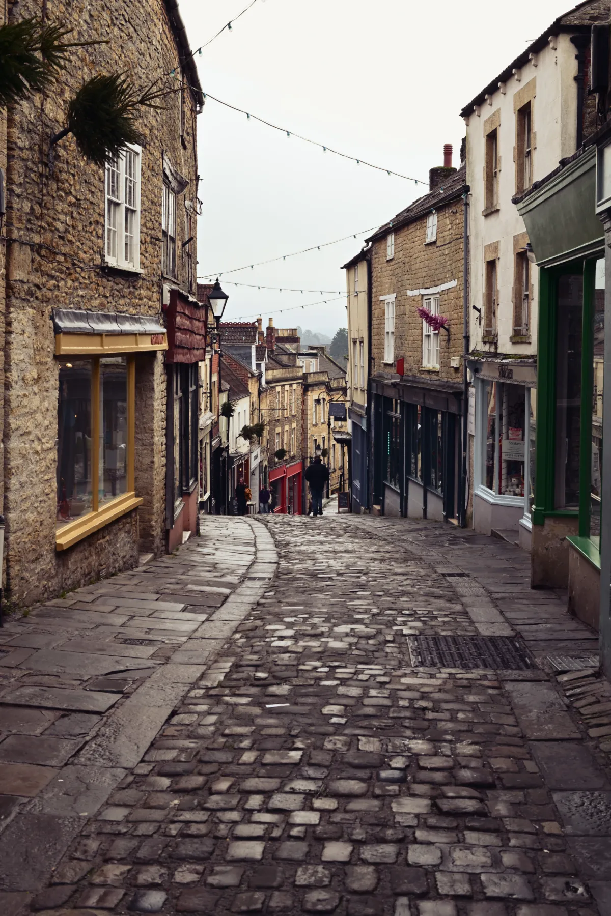 A steep cobbled street lined with shops, strung with overhead lights.
