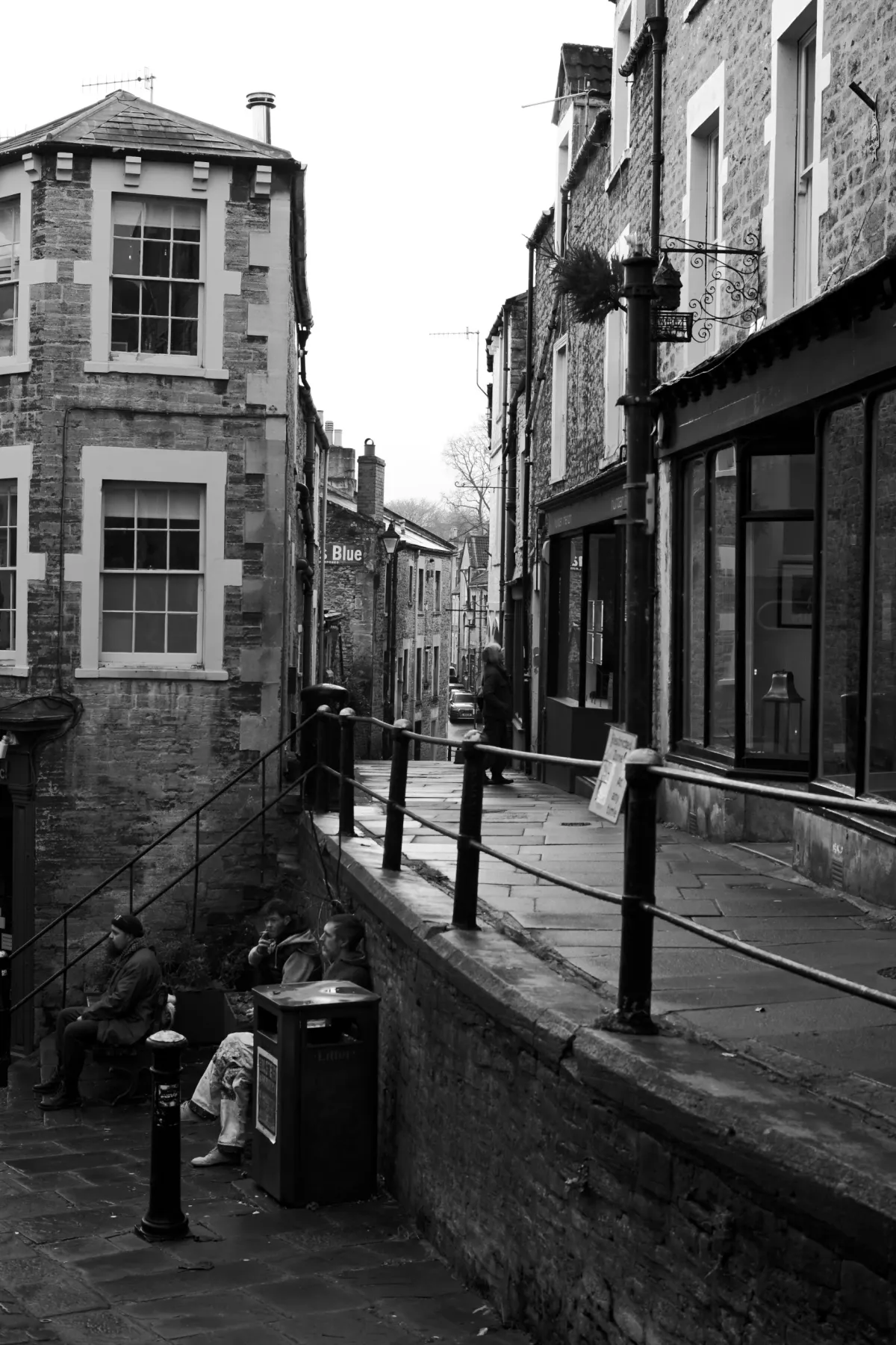 People sitting on a wall above a street, beside a stone building. Black and white.