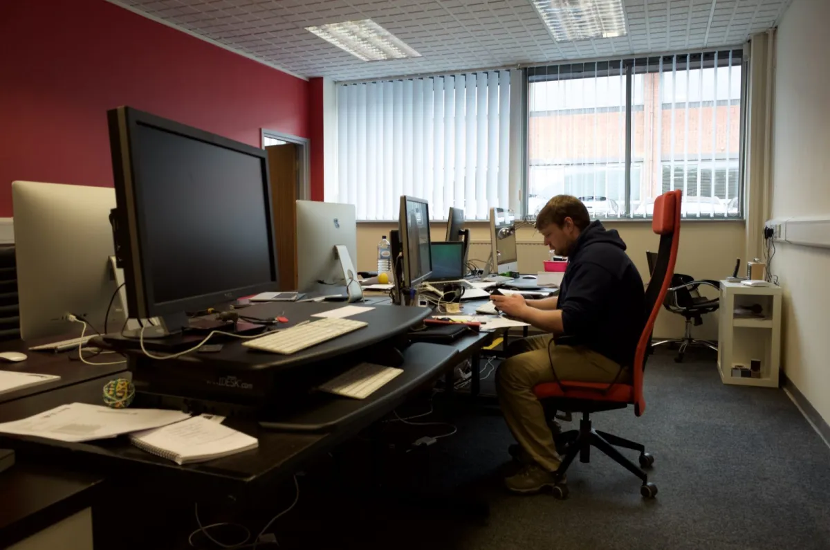 A man working at a desk in an office with a red wall and vertical blinds.