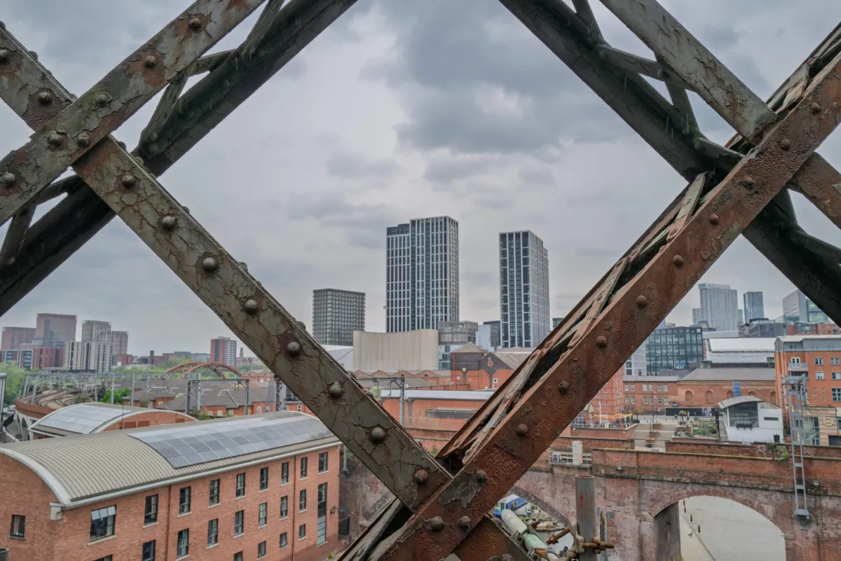 The Manchester skyline framed through the girders of a railway bridge.