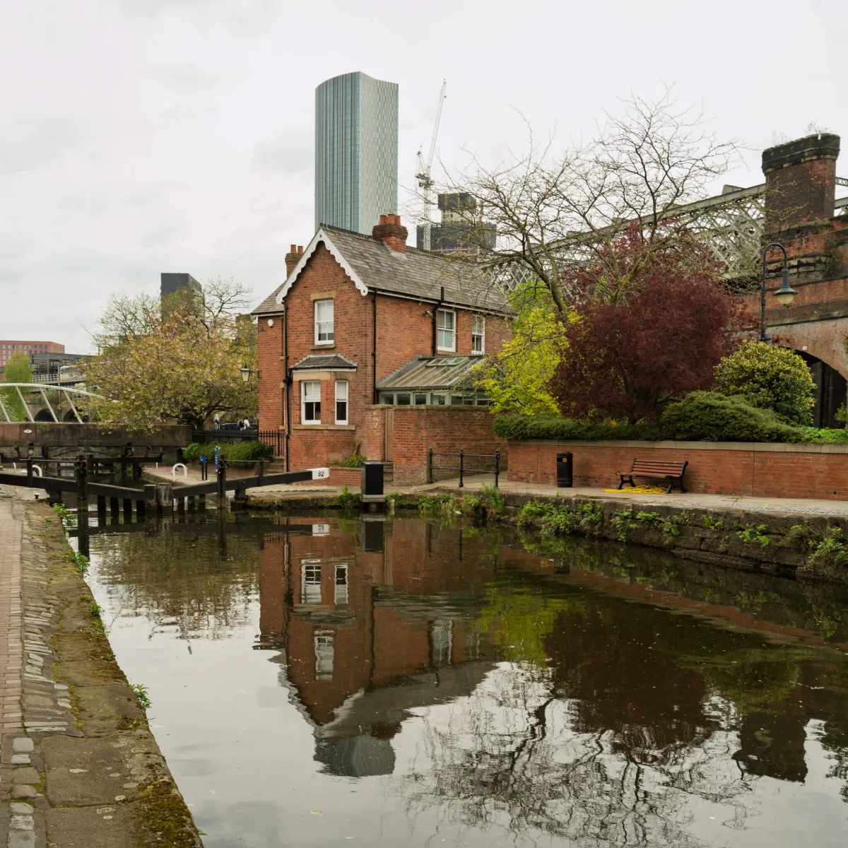 A brick lock keeper's cottage beside a canal, with tower blocks behind.