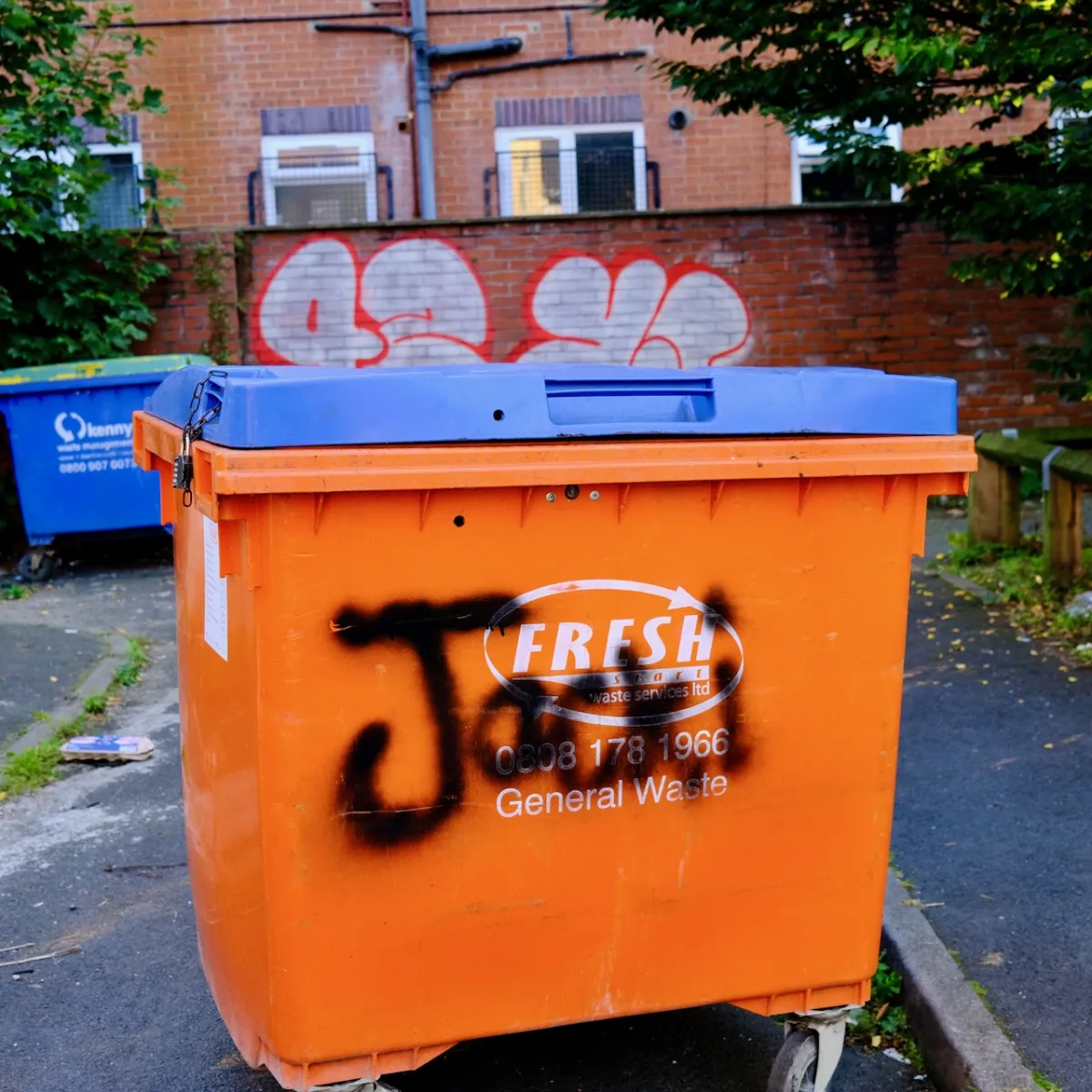 An orange commercial waste bin tagged in black paint, with graffiti on the wall behind.