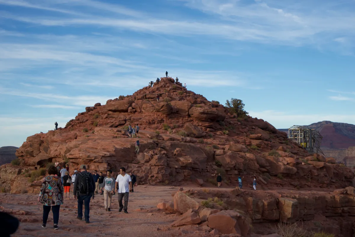 Visitors climbing a rocky red outcrop at the canyon rim under a blue sky.