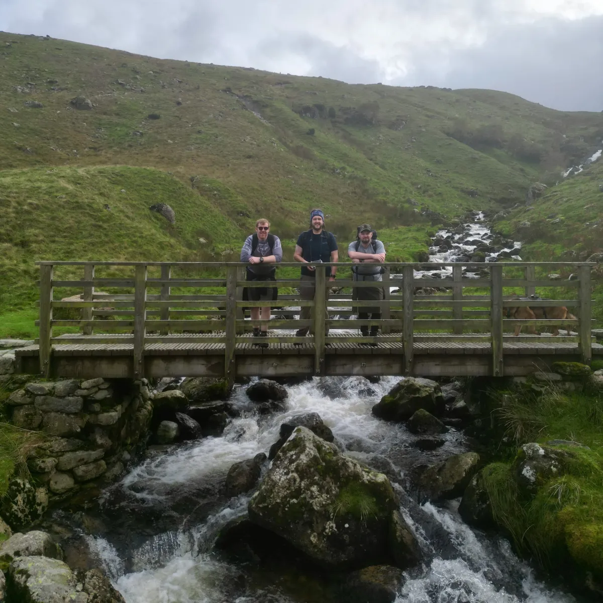 Three walkers standing on a wooden footbridge over a rocky stream, with grassy fellside behind.