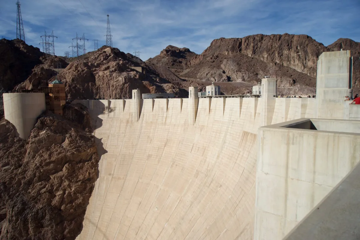 The curved concrete crest of the Hoover Dam, with electricity pylons on the hills behind.