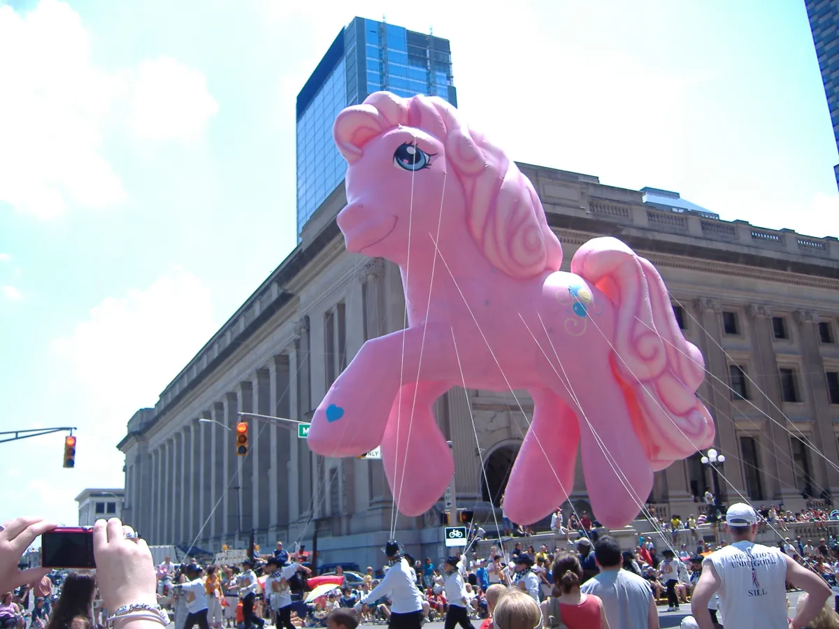 A large pink pony parade balloon held on lines above a crowded street.