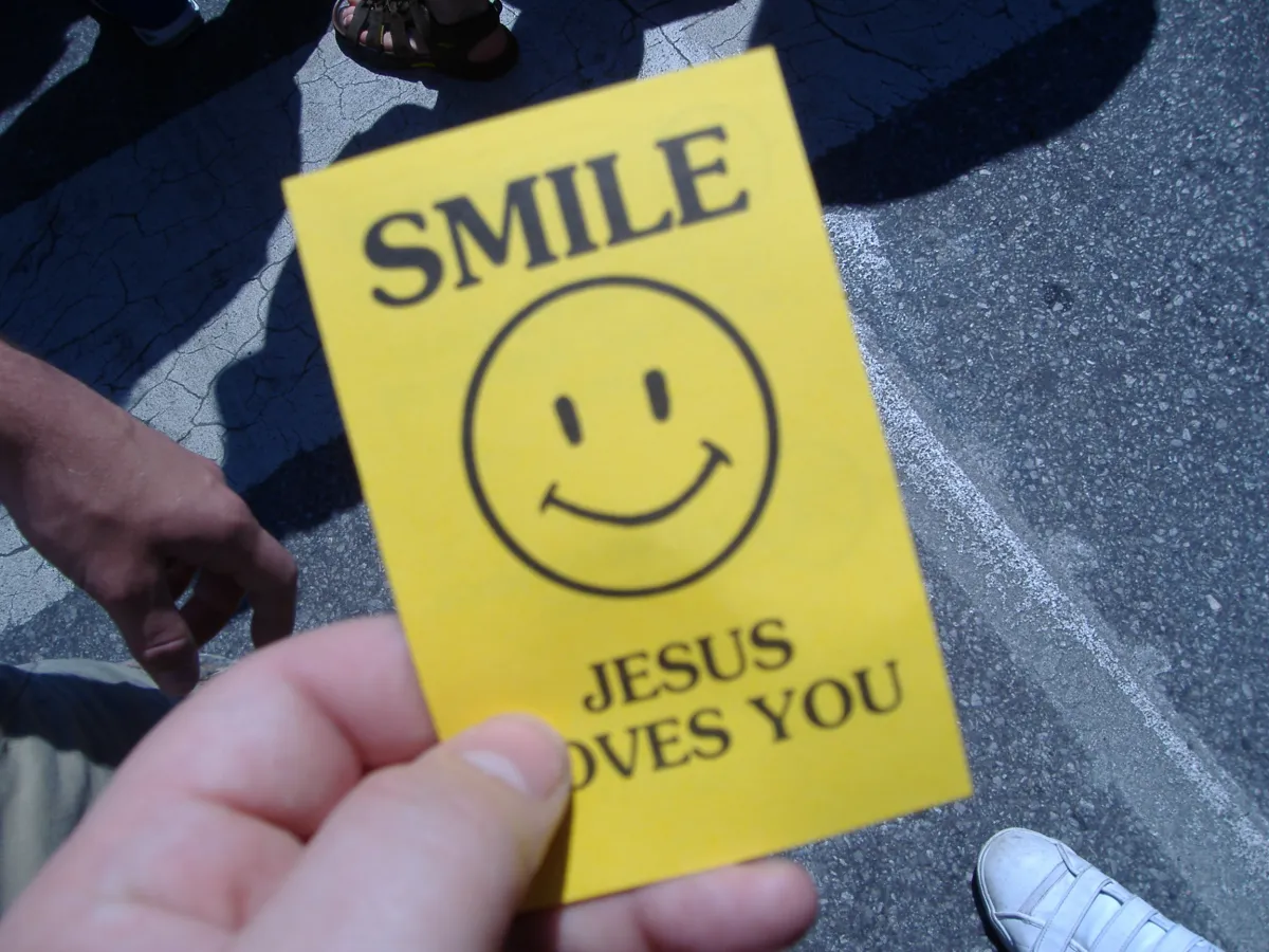 A hand holding a yellow flyer printed with a smiley face and the words Smile, Jesus Loves You.