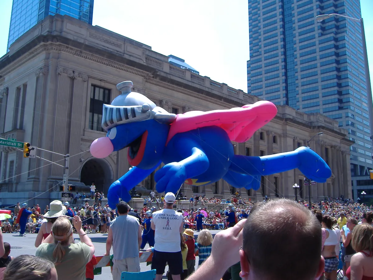 A large blue superhero parade balloon floating between city buildings above a crowd.