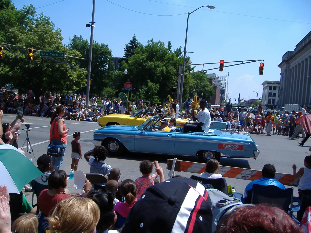 A blue convertible carrying performers along a parade route lined with spectators.