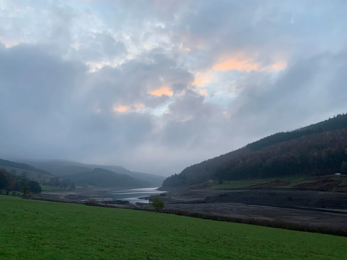 A reservoir at low water level seen across a field, under heavy cloud at dusk.