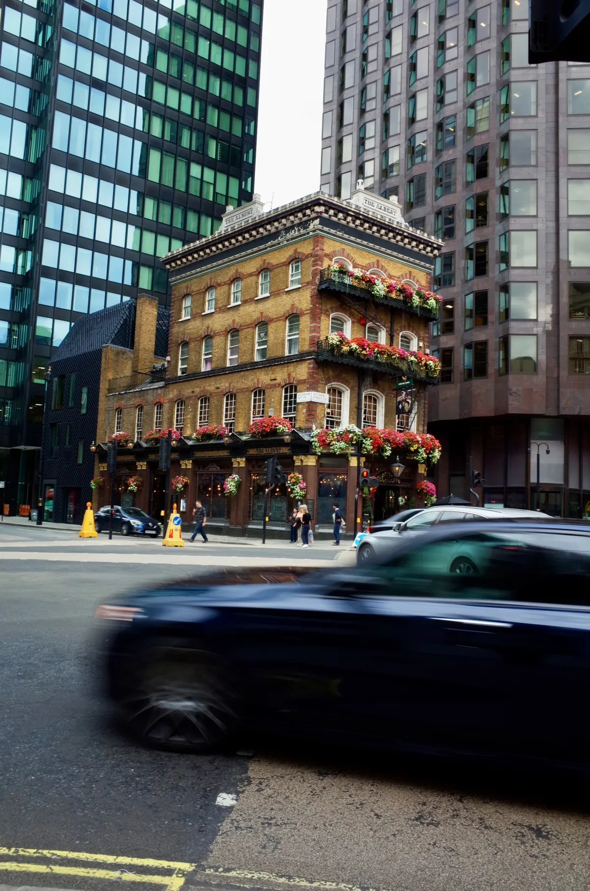 A brick corner pub hung with flower baskets, between glass office blocks, with a car passing.