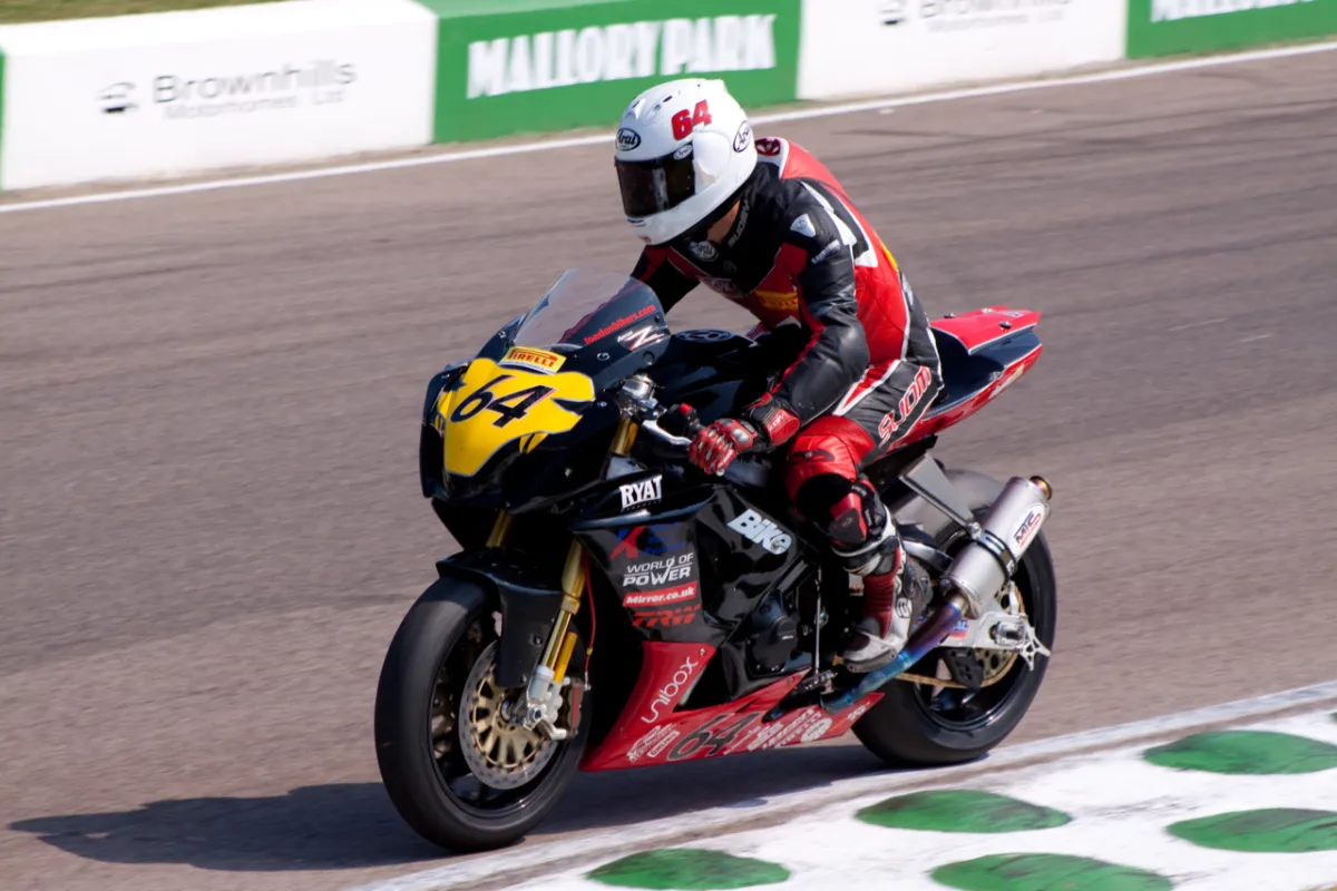 A rider on a black and yellow motorcycle numbered 64, with a Mallory Park hoarding behind.