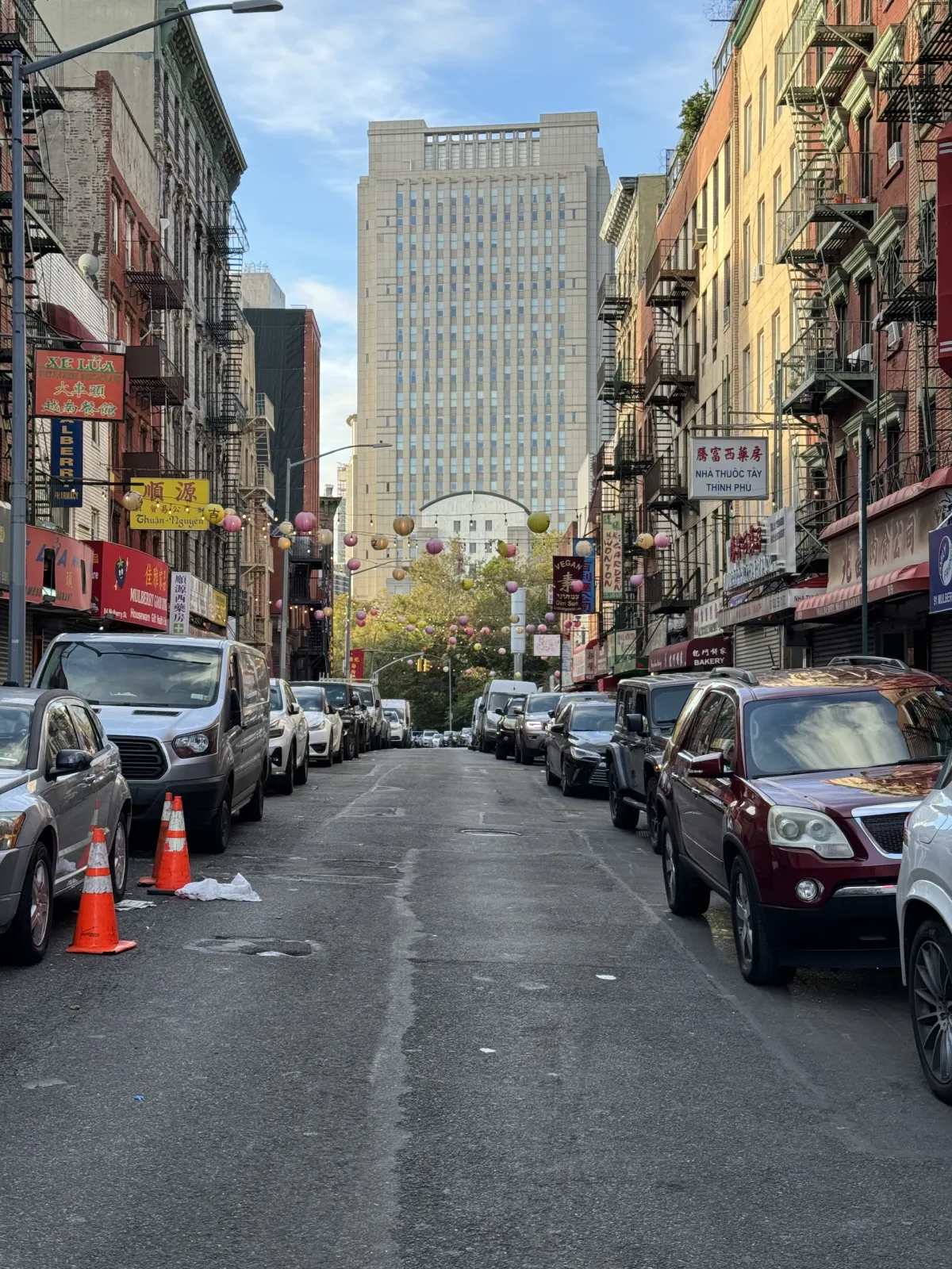 A street in Chinatown lined with parked cars and shop signs, with a tall building at the end.