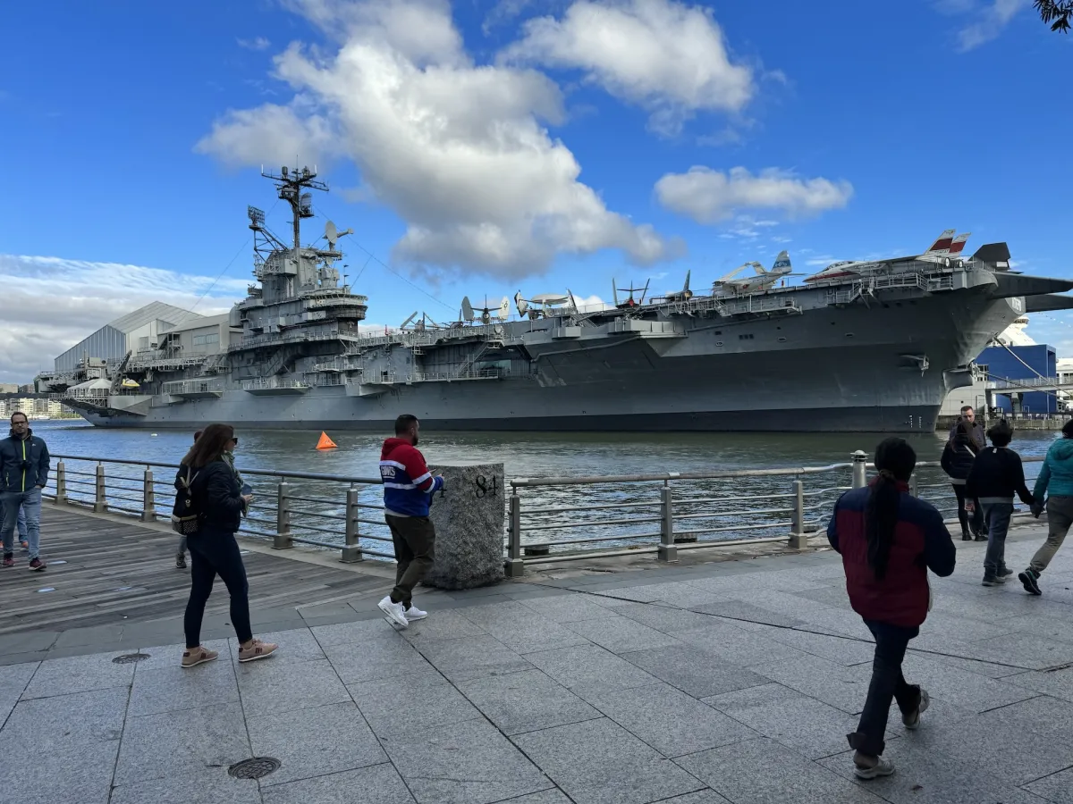 The aircraft carrier Intrepid moored at a pier, with people walking along the waterfront.