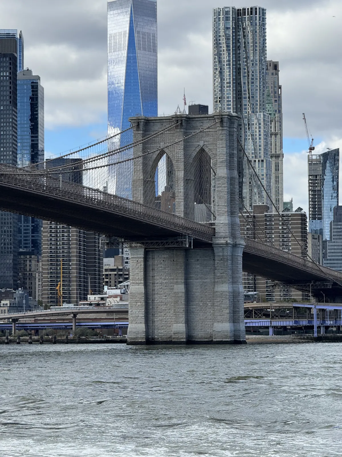 A stone tower of the Brooklyn Bridge seen from the water, with skyscrapers behind.