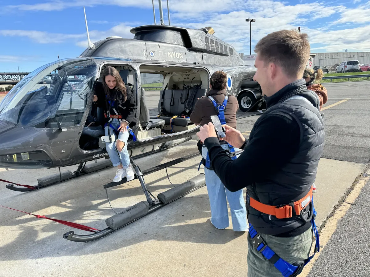 People being fitted with harnesses beside a helicopter on a landing pad.