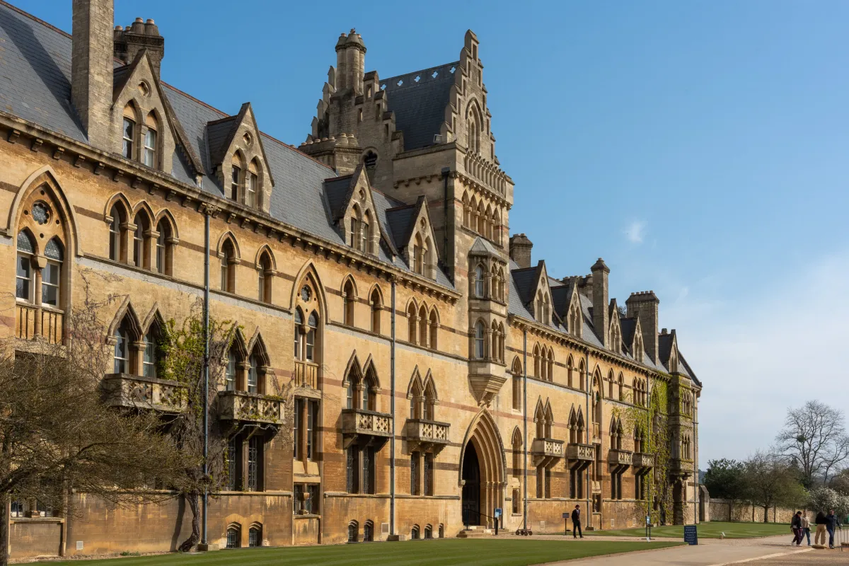 The long gothic frontage of a college building beside a lawn, under a clear sky.