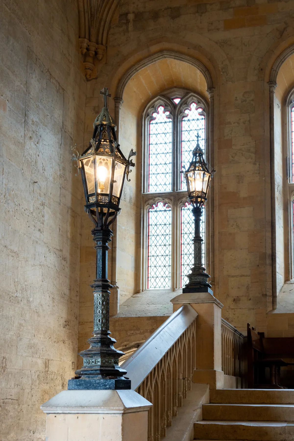 Two ornate iron lamps on stone plinths beside a staircase, below a stained-glass window.