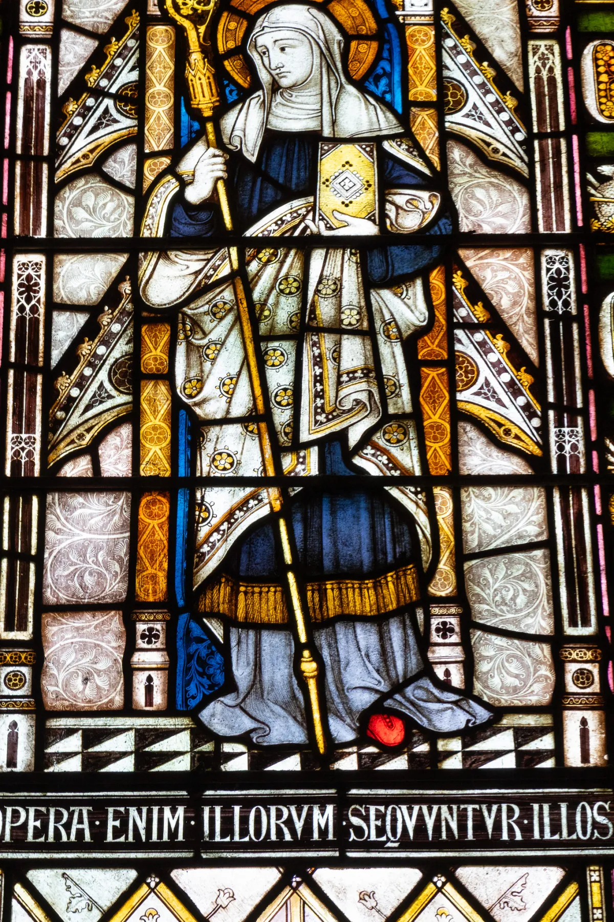 A stained-glass panel showing a robed figure holding a staff, above a Latin inscription.