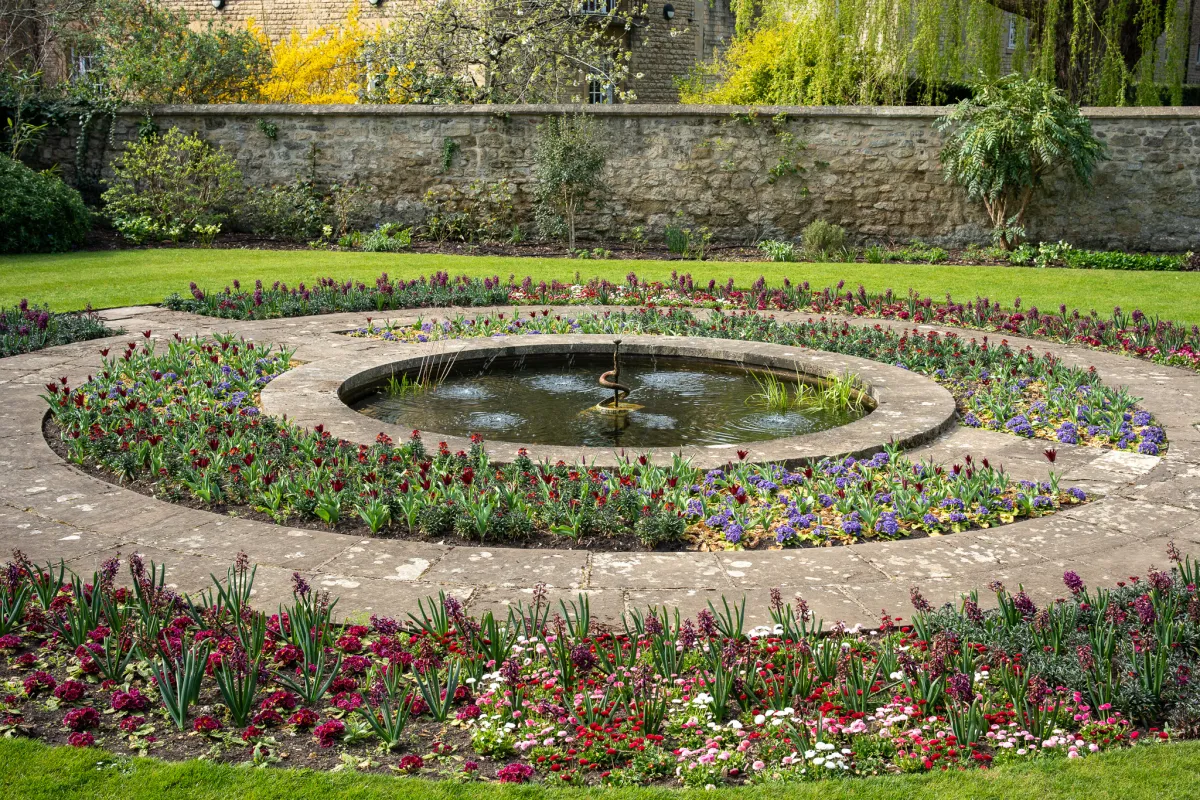 A circular pond and fountain set in a walled garden, ringed by beds of spring flowers.