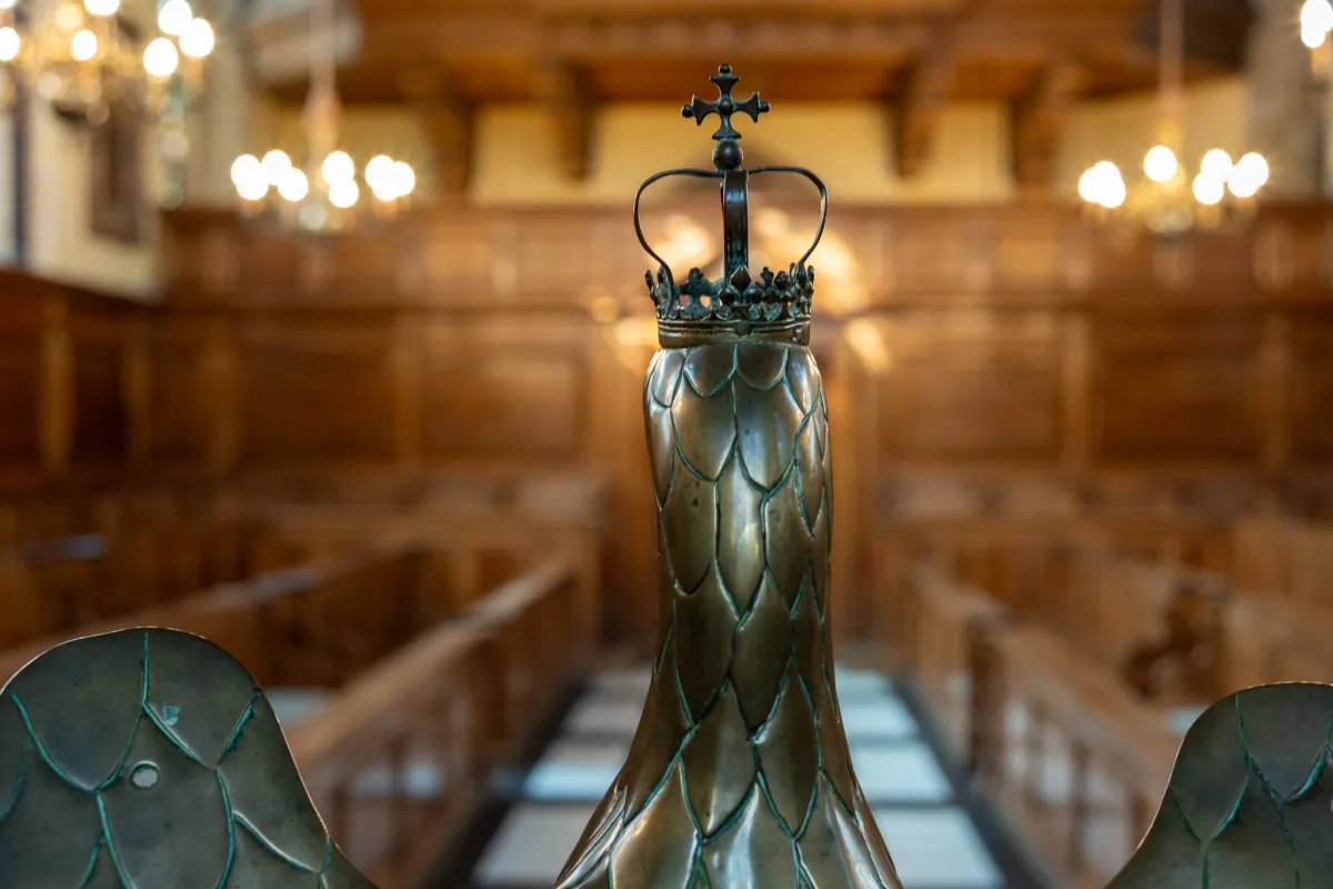A brass finial topped with a crown, in front of tiered wooden benches in a panelled hall.