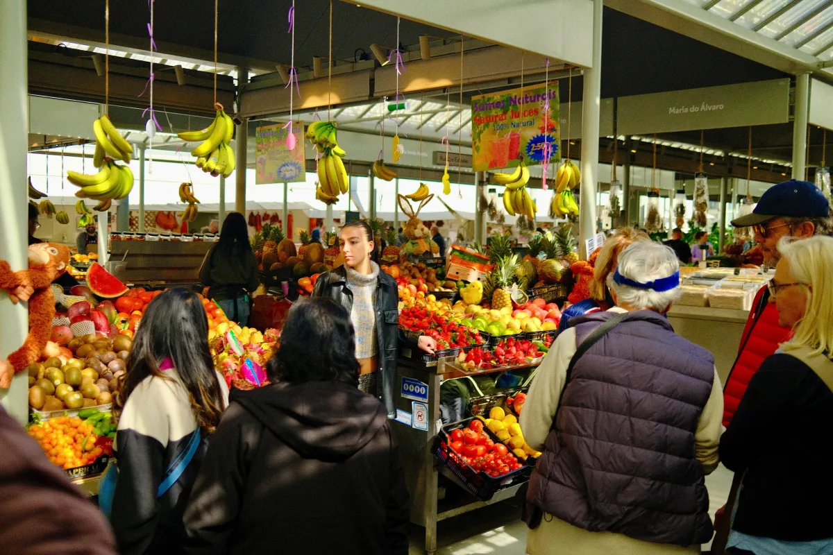 Shoppers browsing a covered fruit market, bunches of bananas hanging above the stalls.