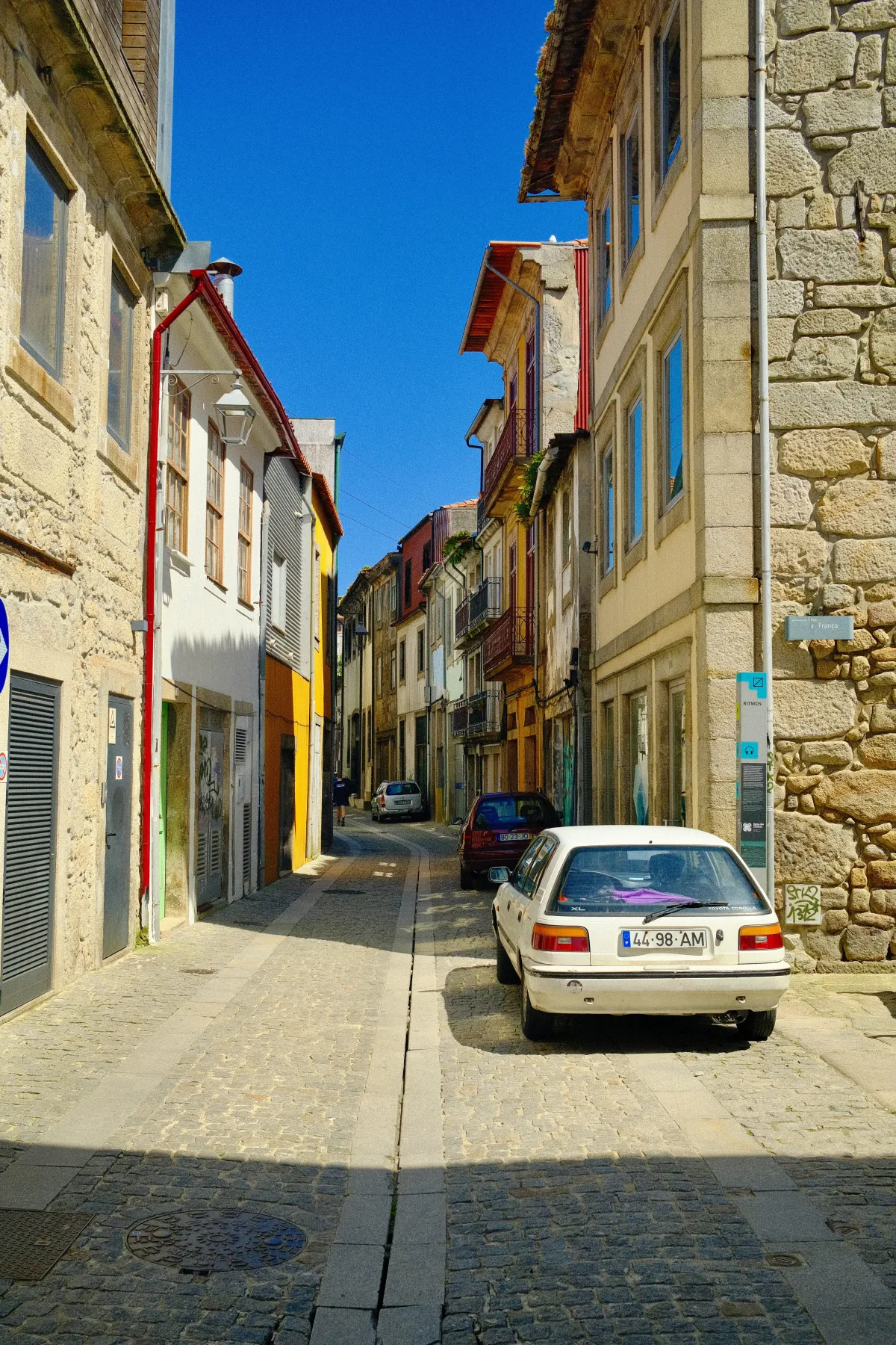 A narrow cobbled street between stone buildings, with a white car parked at the kerb.