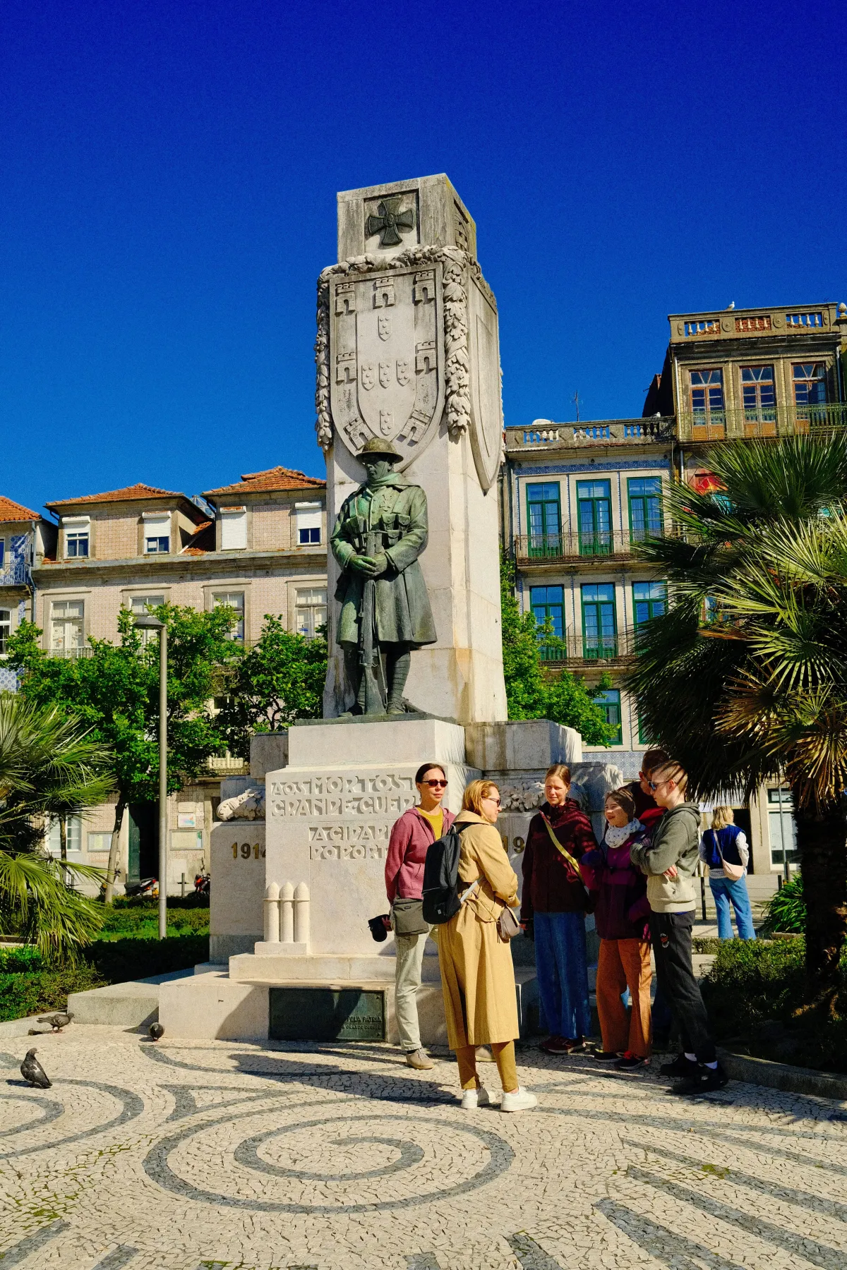 A stone war memorial topped by a soldier, with visitors gathered at its base on patterned paving.