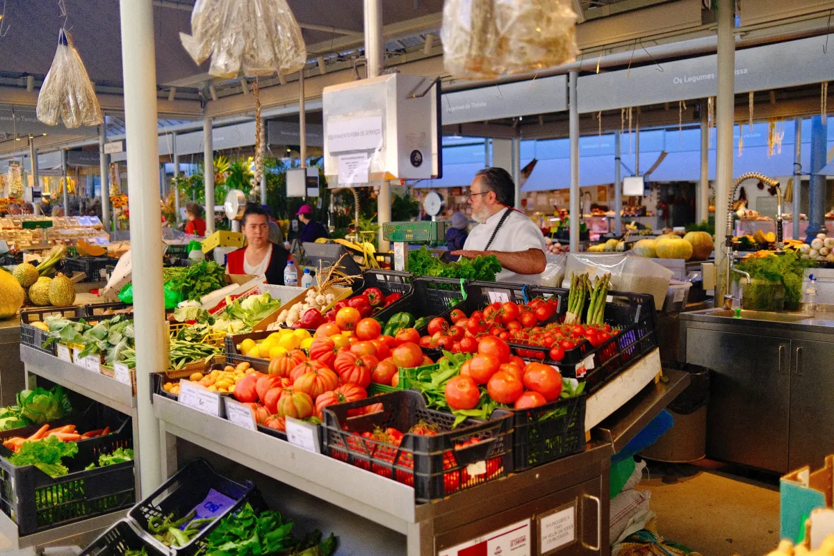 A greengrocer's stall in a covered market, crates of tomatoes and greens stacked at the front.