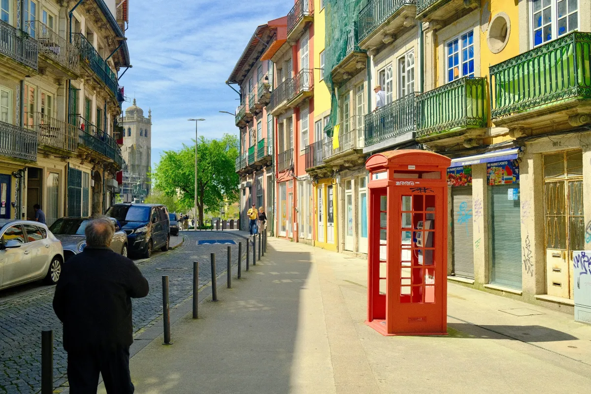 A red telephone box on a street lined with tiled buildings and green wrought-iron balconies.