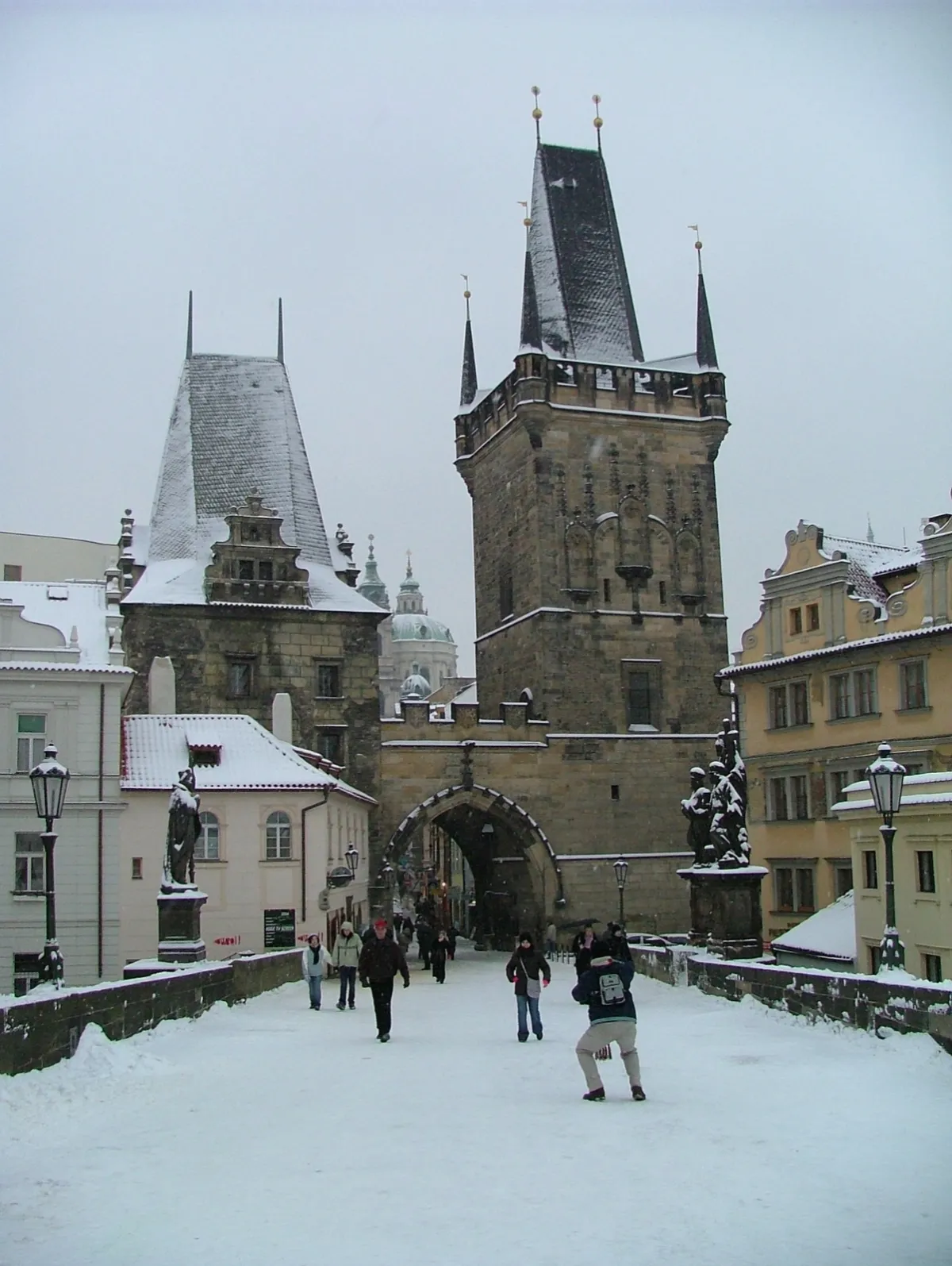 A snow-covered bridge leading to a gothic stone gate tower, with people walking across.