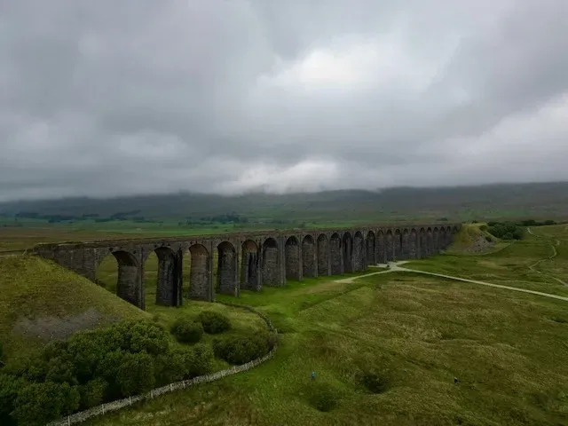 Aerial view of the Ribblehead Viaduct curving across green moorland, its stone arches stretching towards hills shrouded in low grey cloud