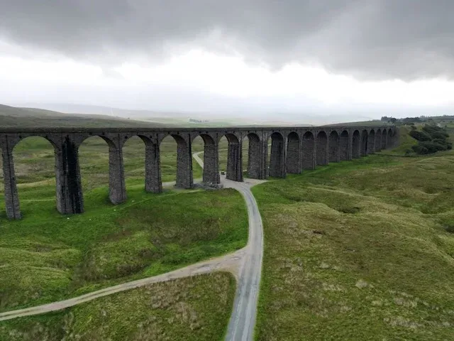 Drone shot looking along the arches of the Ribblehead Viaduct as a narrow lane passes beneath them, under a heavy overcast sky