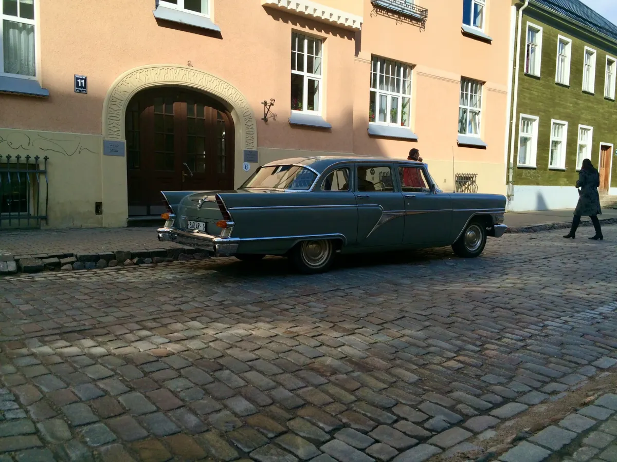 A vintage grey saloon car parked on a cobbled street beside a pink building.