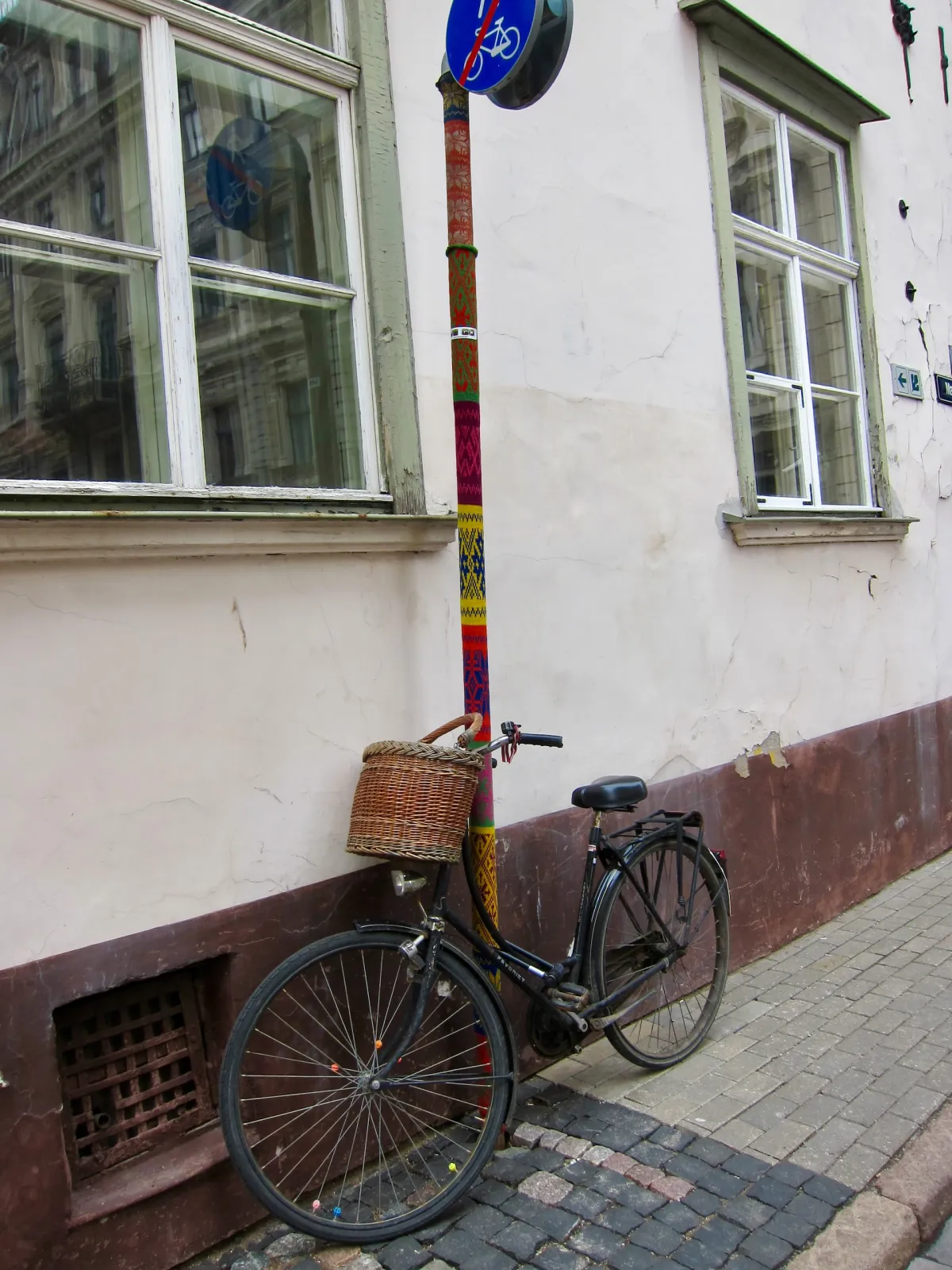 A bicycle with a wicker basket leaning against a lamp post wrapped in knitted yarn.