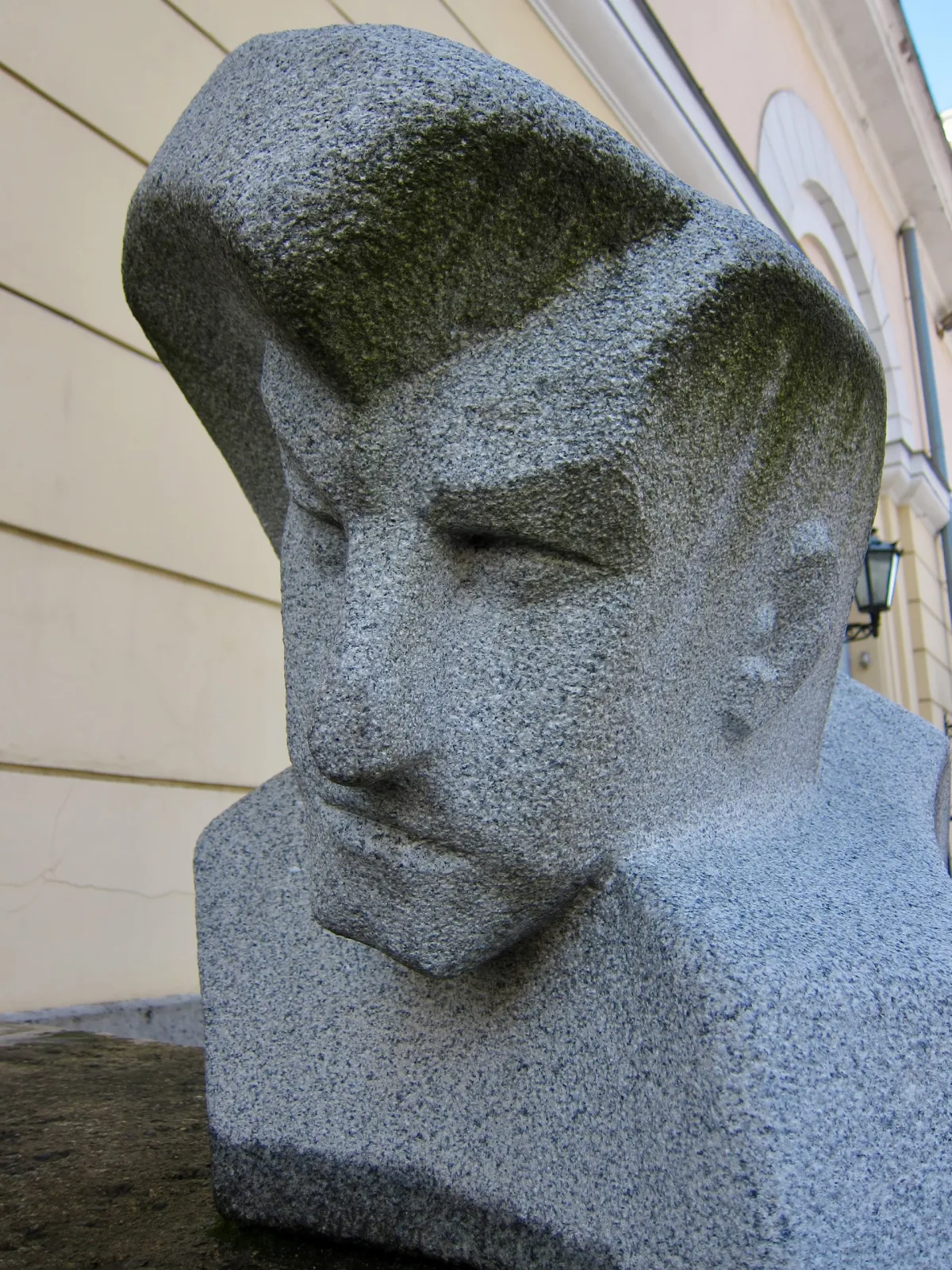 A granite bust of a head with a swept-back fringe, on a pavement outside a building.