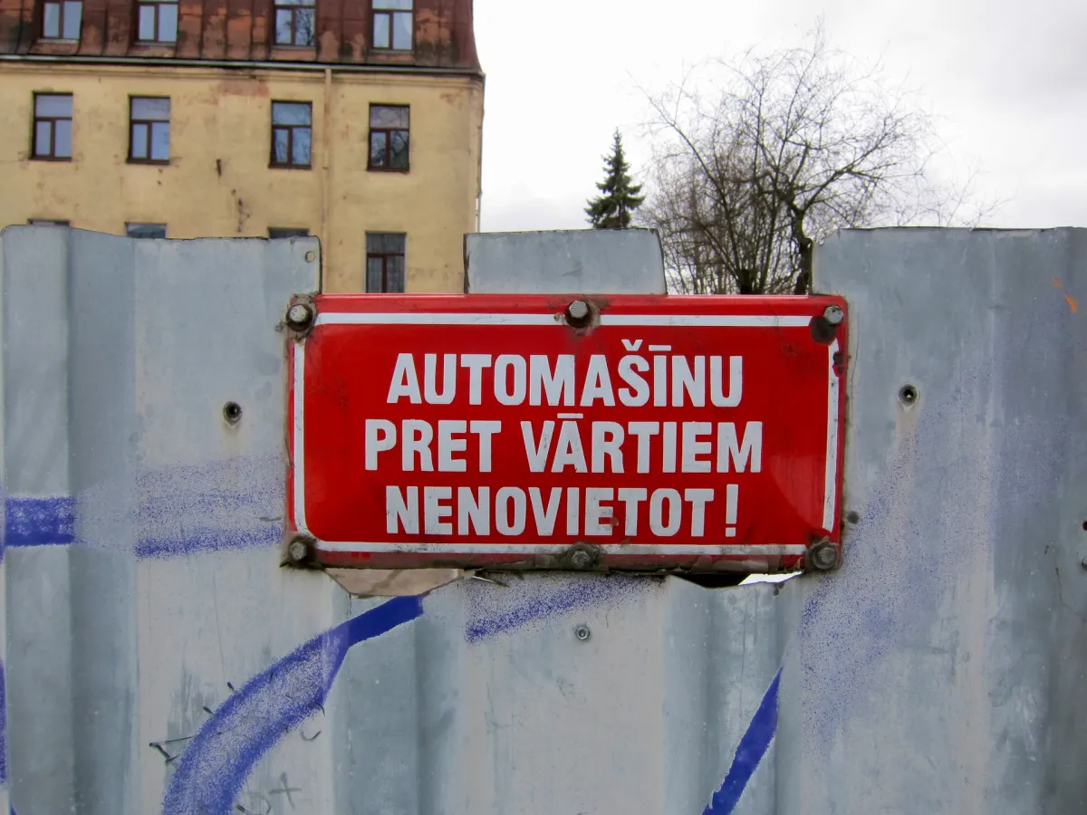 A red enamel sign in Latvian fixed to a concrete hoarding, with a derelict building behind.