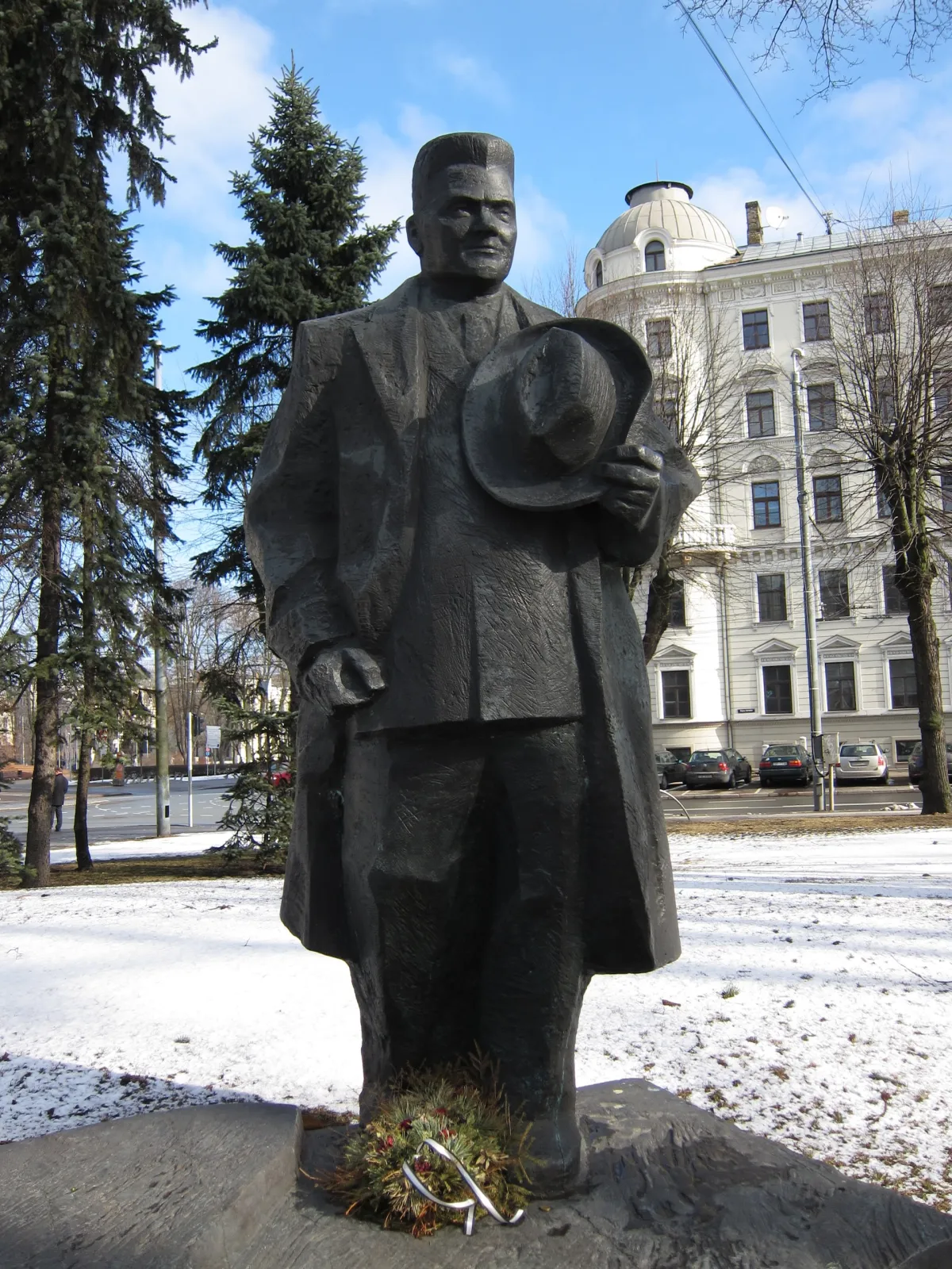 A bronze statue of a man in a long coat holding a hat, in a snow-covered park.